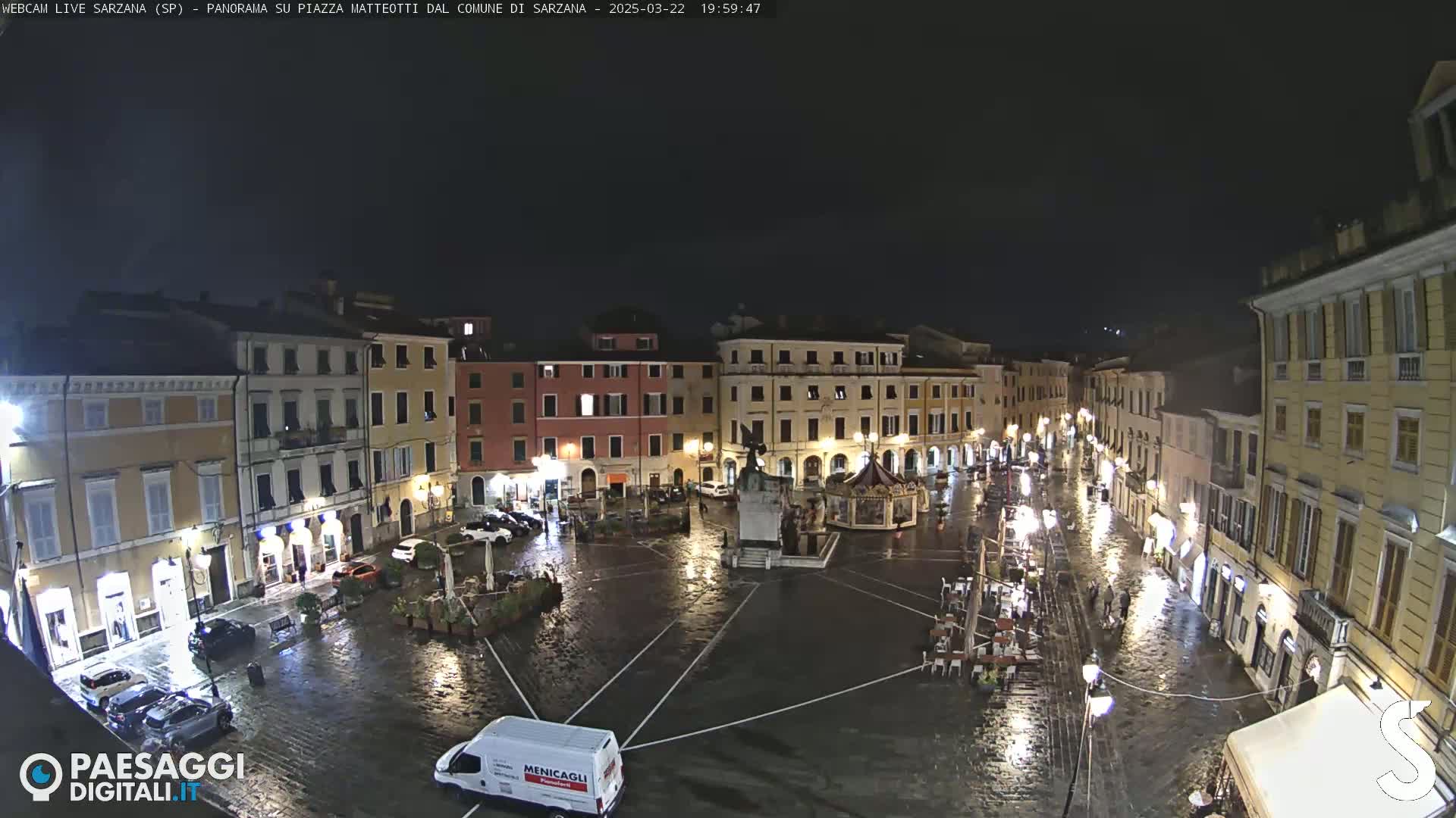 A rain-slicked, nighttime town square is surrounded by illuminated buildings, with a few cars and people visible.