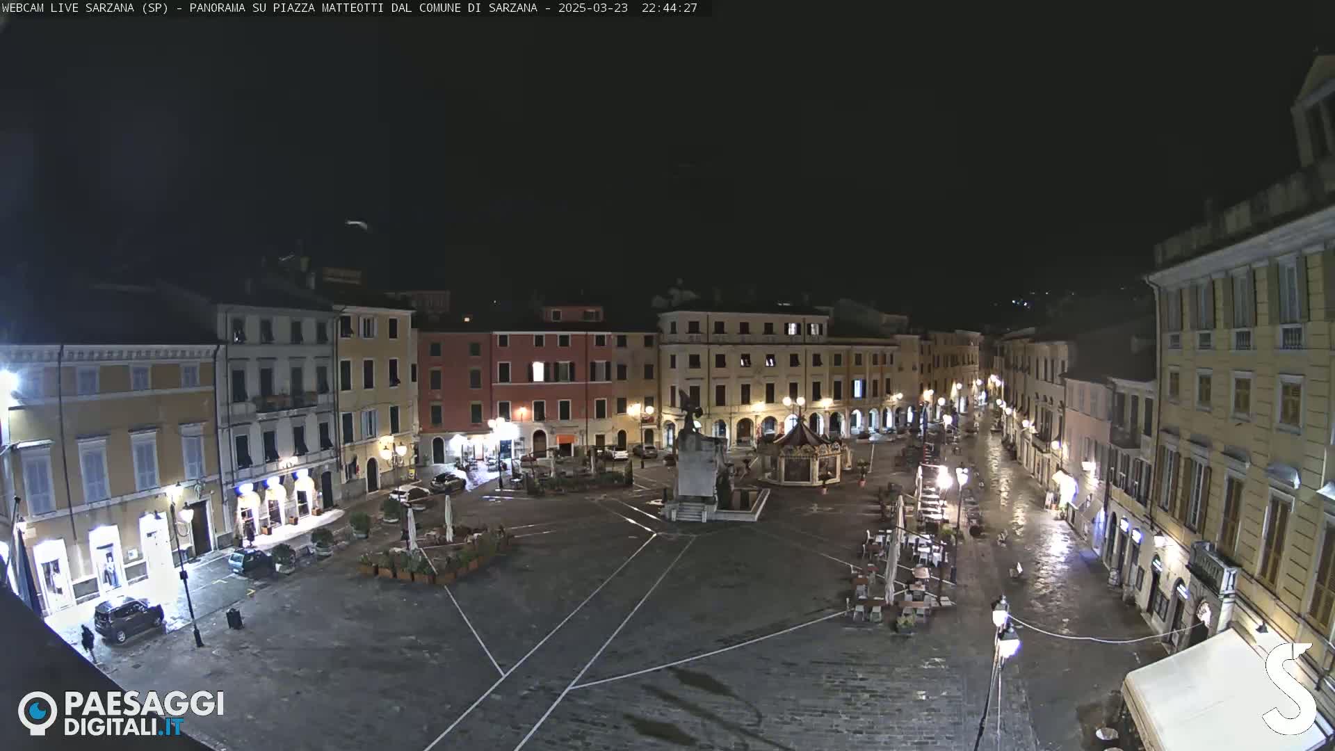 A nighttime view of a town square, wet from recent rain, with several buildings illuminated and sparsely occupied outdoor seating areas.