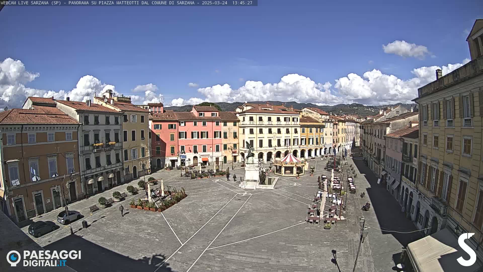 A mostly sunny day reveals a large town square surrounded by historic buildings, with a few people and outdoor seating visible.