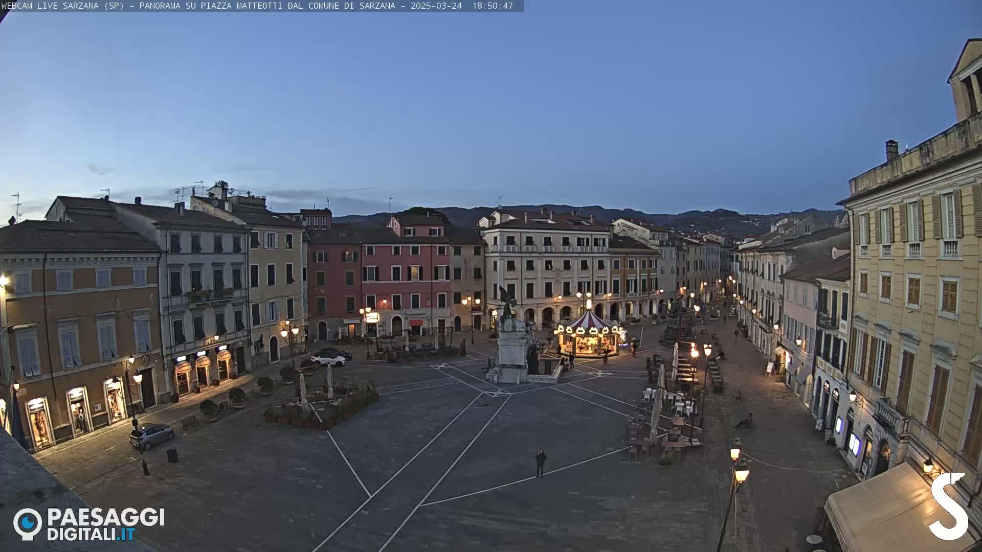A town square at twilight, illuminated by streetlights, features multi-story buildings lining its perimeter, a carousel, and a central monument.