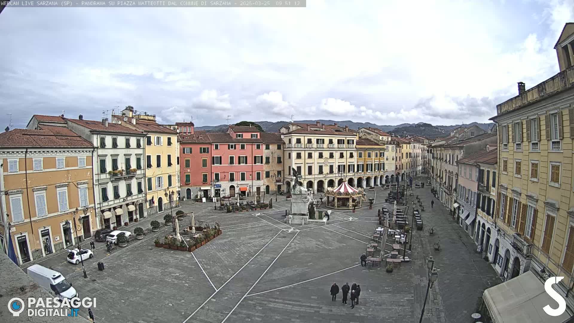 A mostly empty town square in a European town, under a partly cloudy sky, features multi-story buildings surrounding it with a few people and some outdoor seating visible.