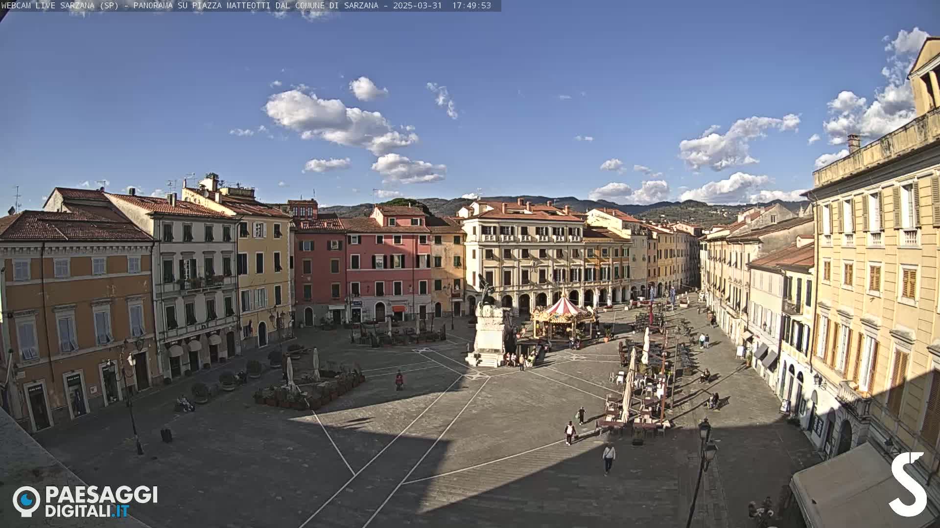 A large, mostly empty town square with several buildings surrounding it, under a mostly sunny sky with some clouds.