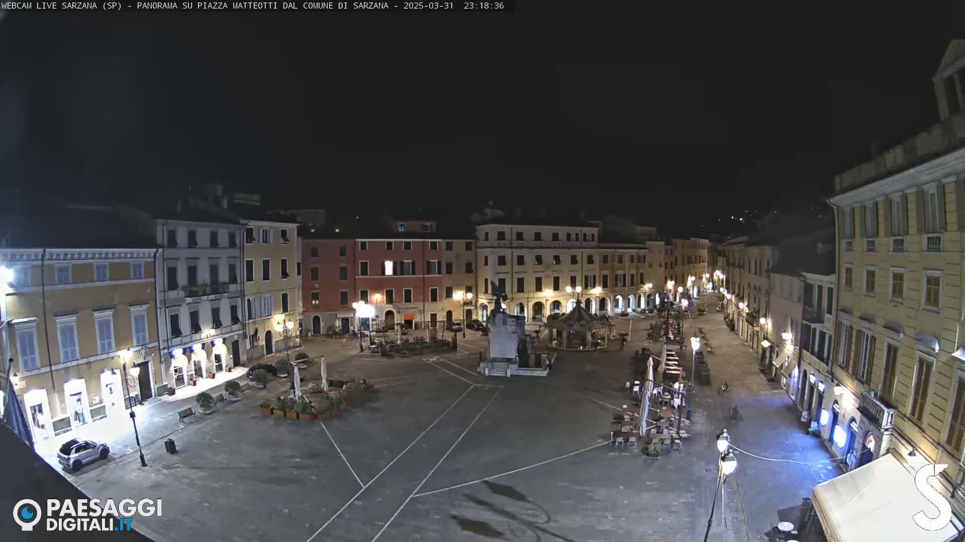 A nighttime aerial view of a mostly empty town square, surrounded by buildings with illuminated windows and outdoor seating areas.