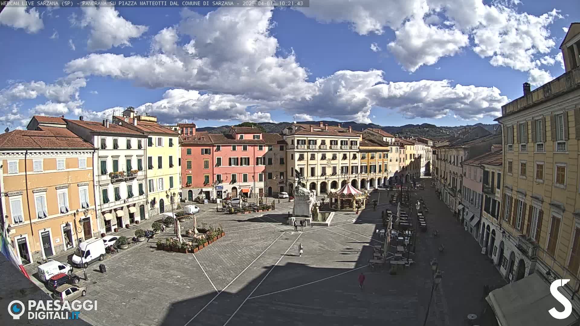 A partly cloudy day overlooks a large town square surrounded by multi-story buildings, with a few people and some outdoor seating visible.