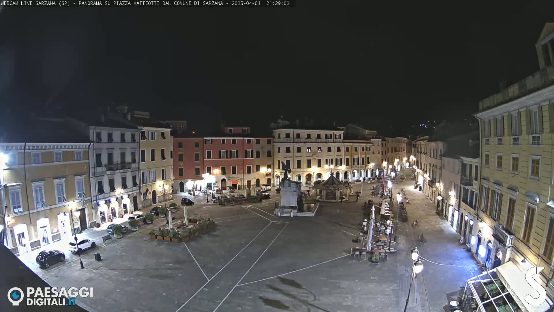 A mostly empty town square at night, illuminated by streetlights, is surrounded by multi-story buildings.
