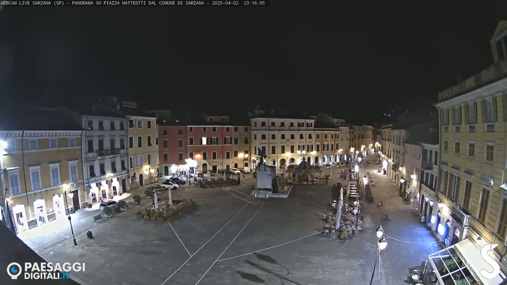 A nighttime view of a town square, mostly empty except for a few cars and outdoor seating areas at cafes, surrounded by multi-story buildings.