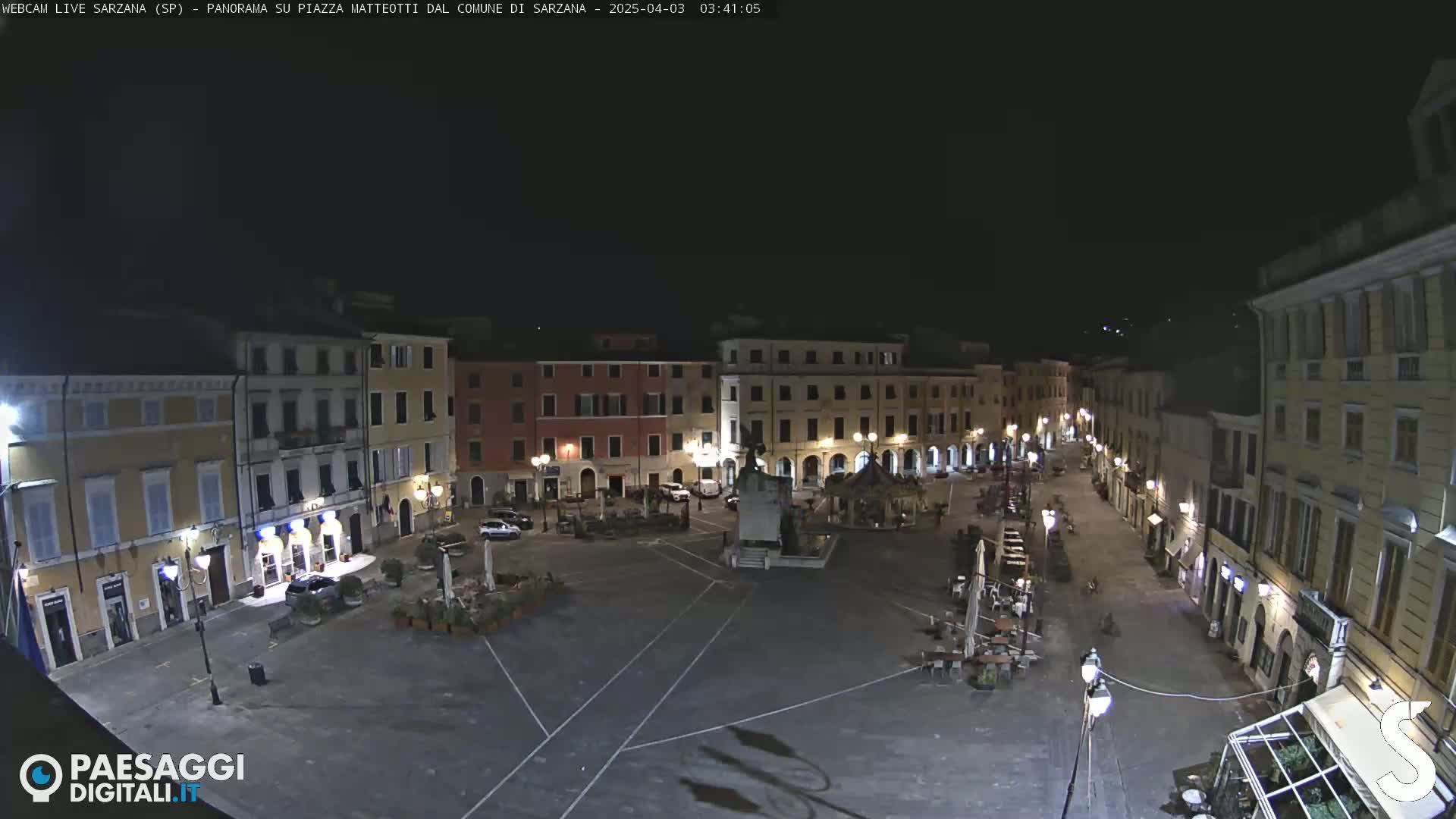 A nighttime, high-angle view of a mostly empty town square, surrounded by illuminated buildings.