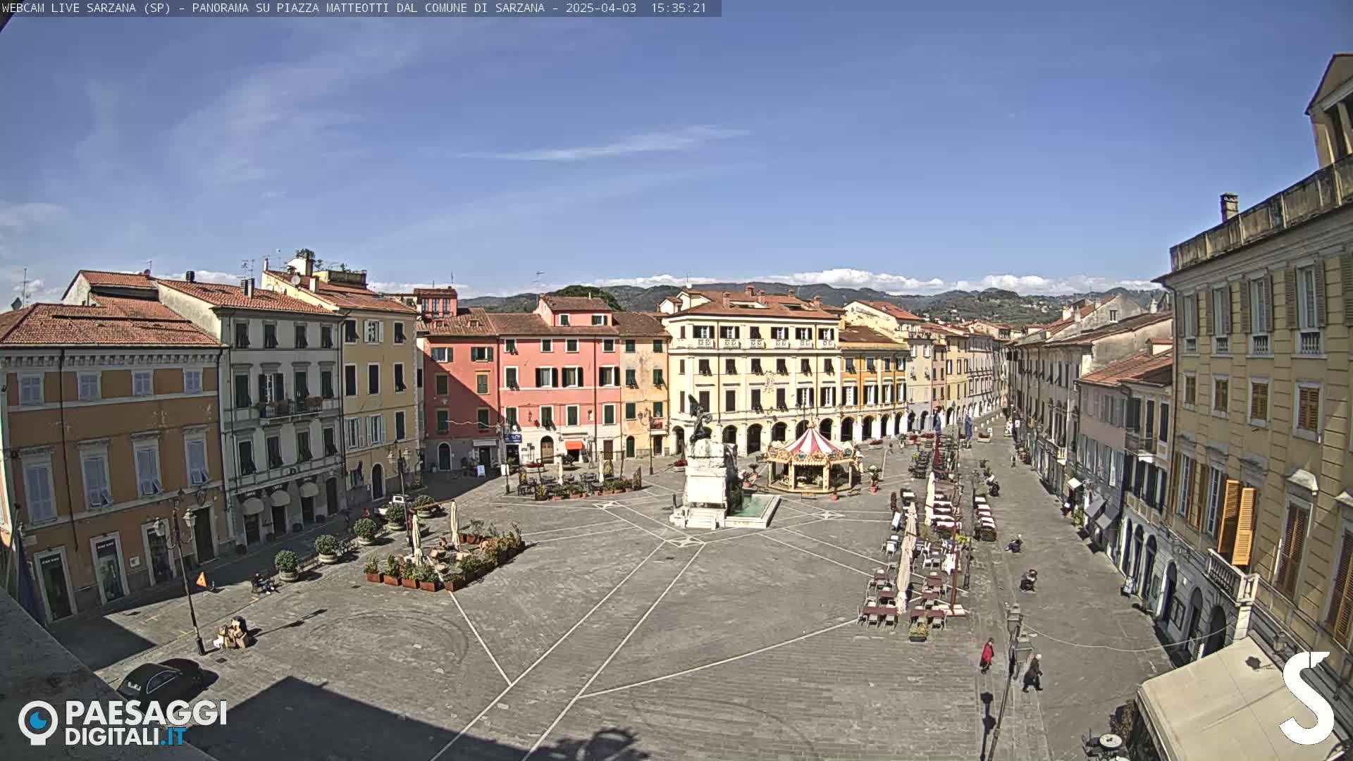 A large, mostly empty town square with surrounding buildings under a clear, sunny sky.