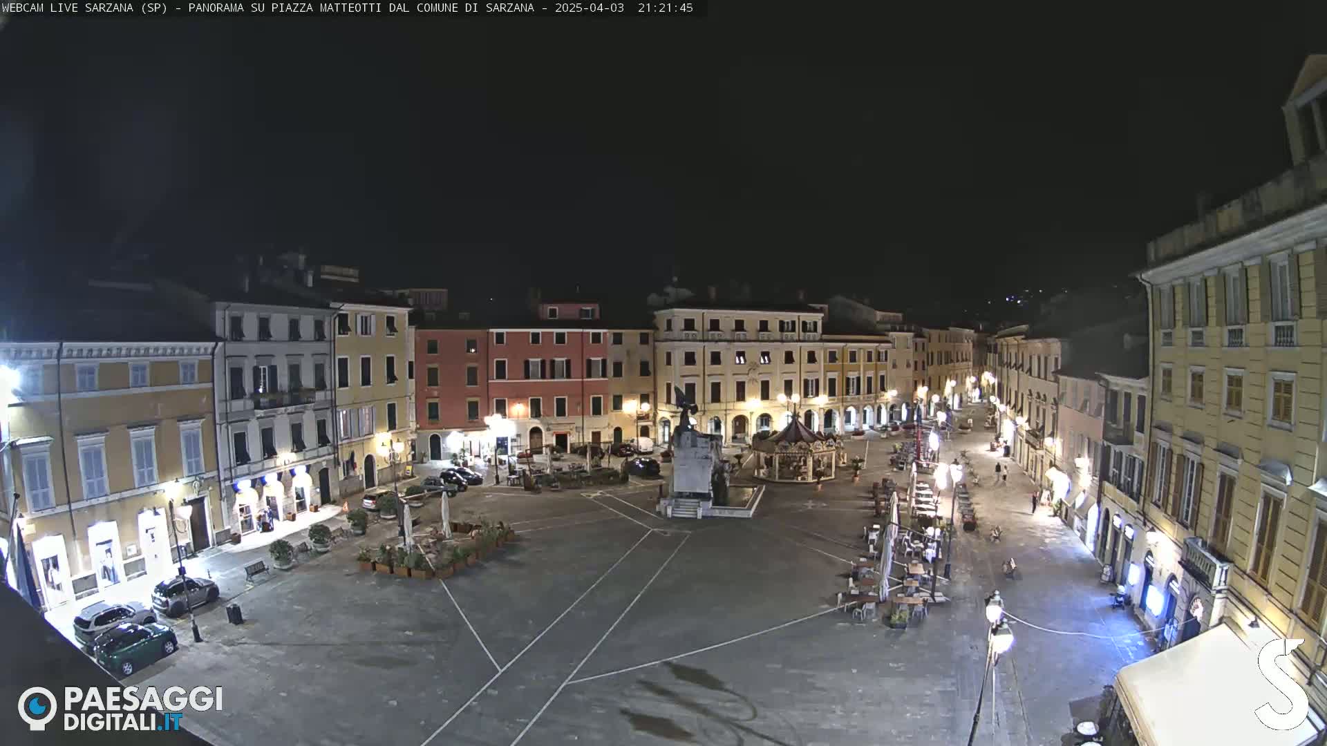 A nighttime view of a town square, surrounded by buildings with illuminated windows and outdoor seating areas.