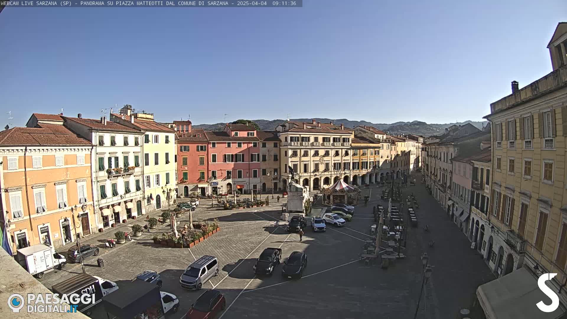 A sunny town square, surrounded by multi-story buildings, features several parked cars and outdoor seating areas.