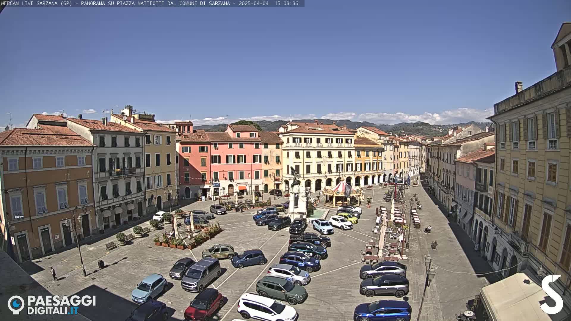 A sunny day reveals a town square filled with parked cars, outdoor seating, and historic buildings under a clear blue sky.