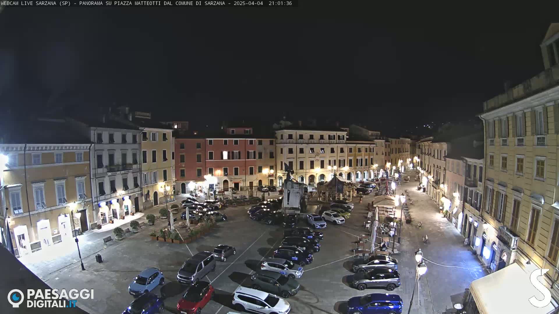 A nighttime view of a town square with parked cars, surrounded by buildings lit with streetlights.
