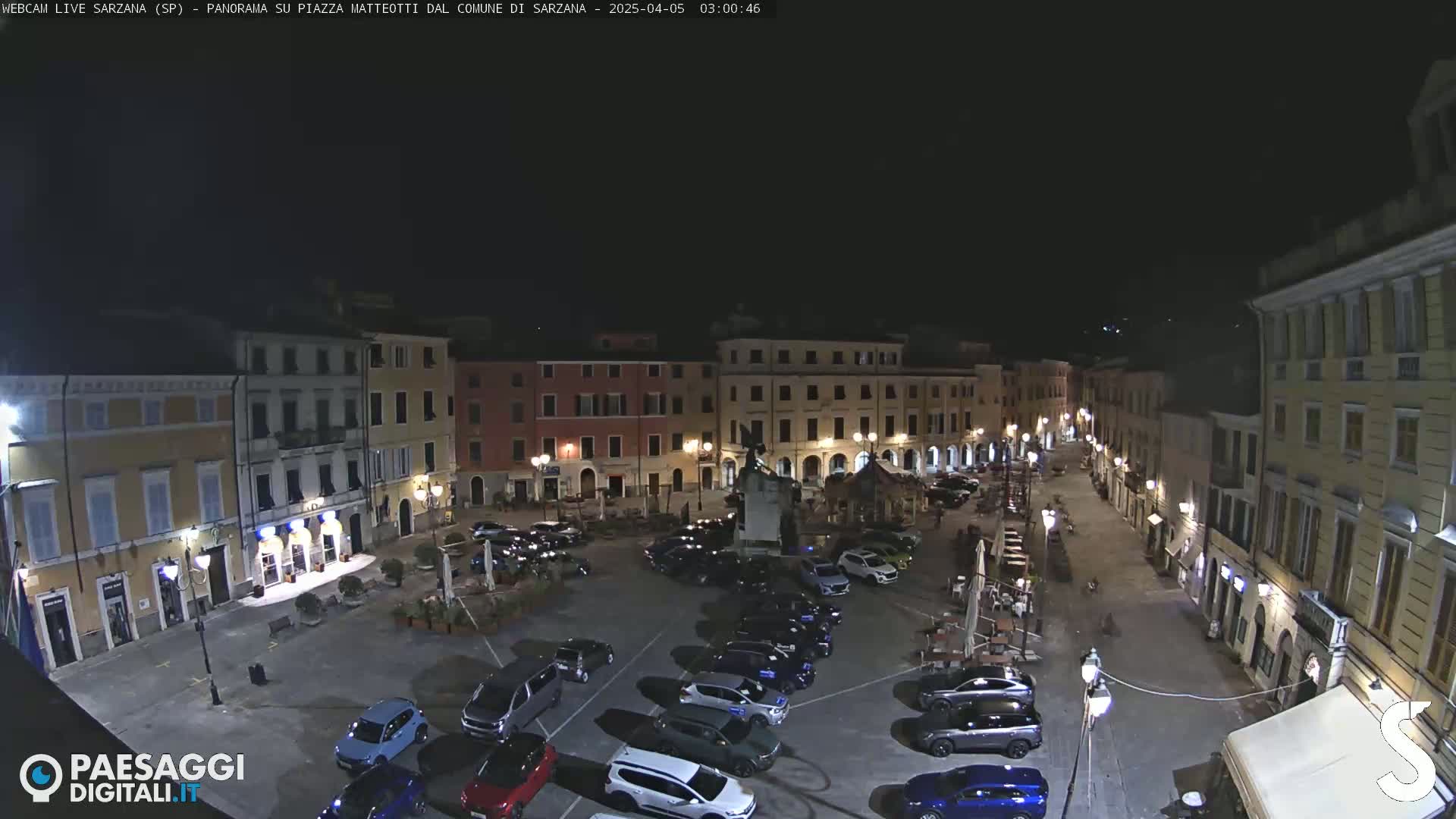 A nighttime aerial view of a town square with parked cars and buildings illuminated under a clear, dark sky.