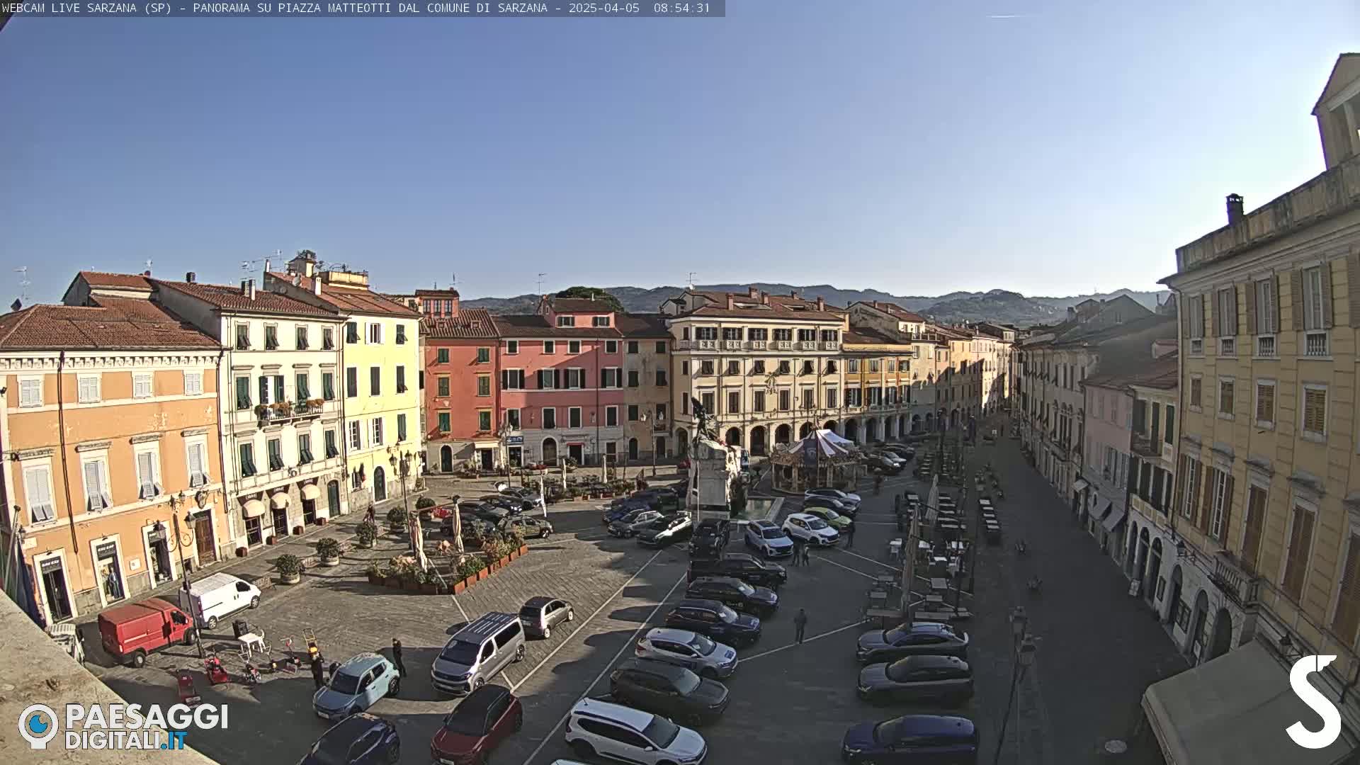 A sunny day in a town square filled with parked cars and several buildings surrounding the open space.