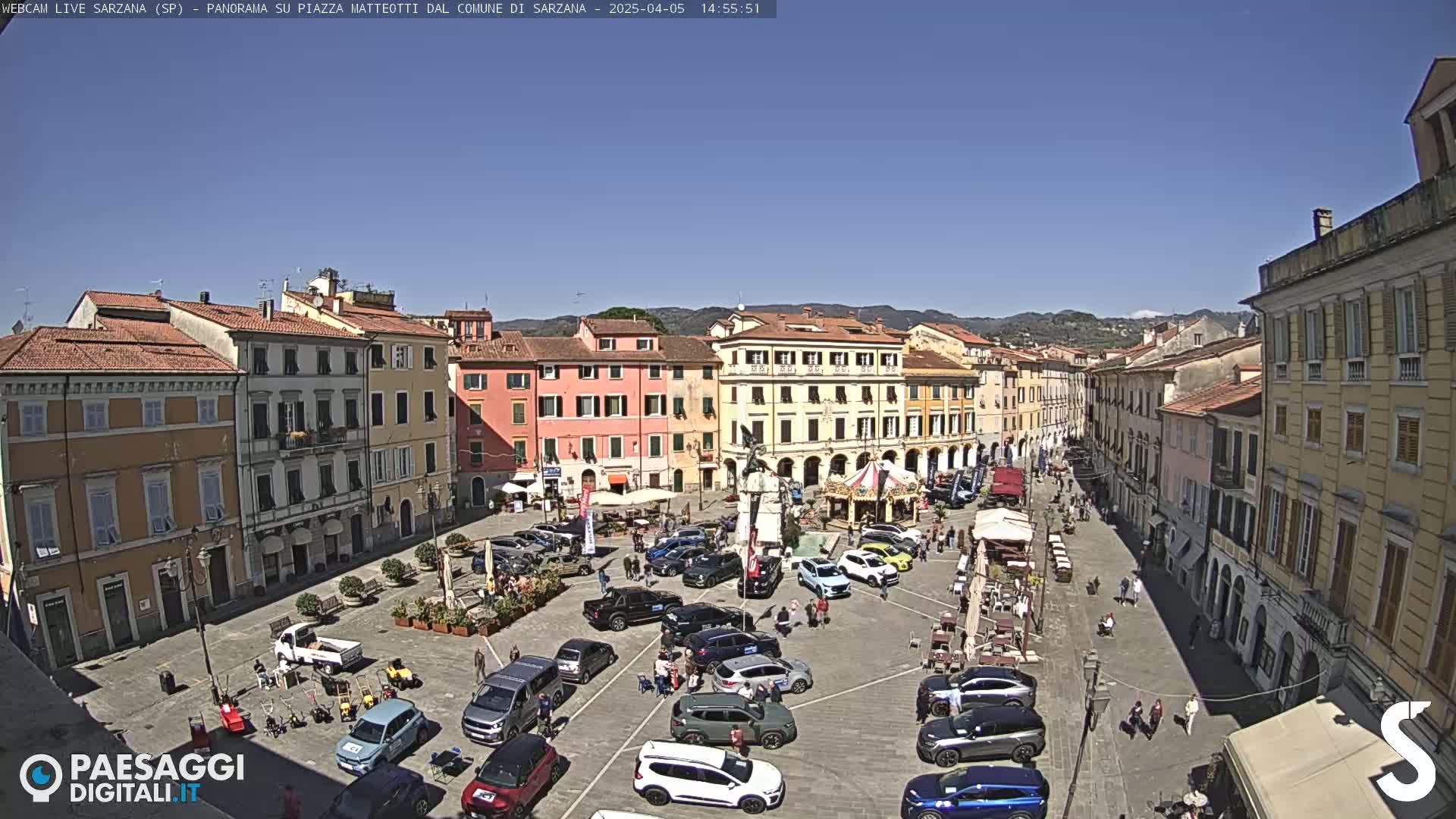 A sunny town square filled with parked cars, outdoor seating, and people walking, surrounded by multi-story buildings.