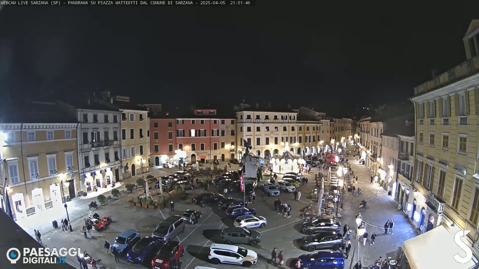 A nighttime, aerial view of a town square filled with parked cars and people, surrounded by illuminated buildings.
