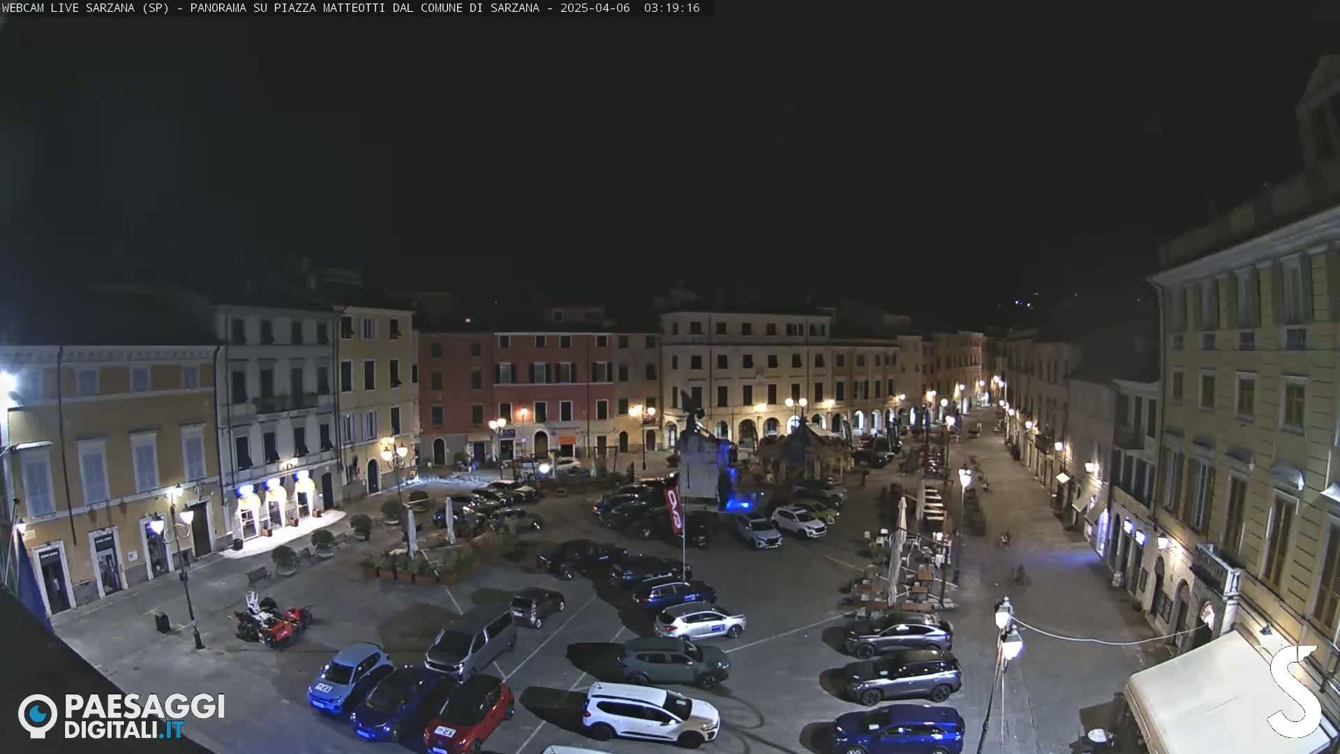 A nighttime, aerial view of a town square filled with parked cars, surrounded by buildings with illuminated windows and streetlights.