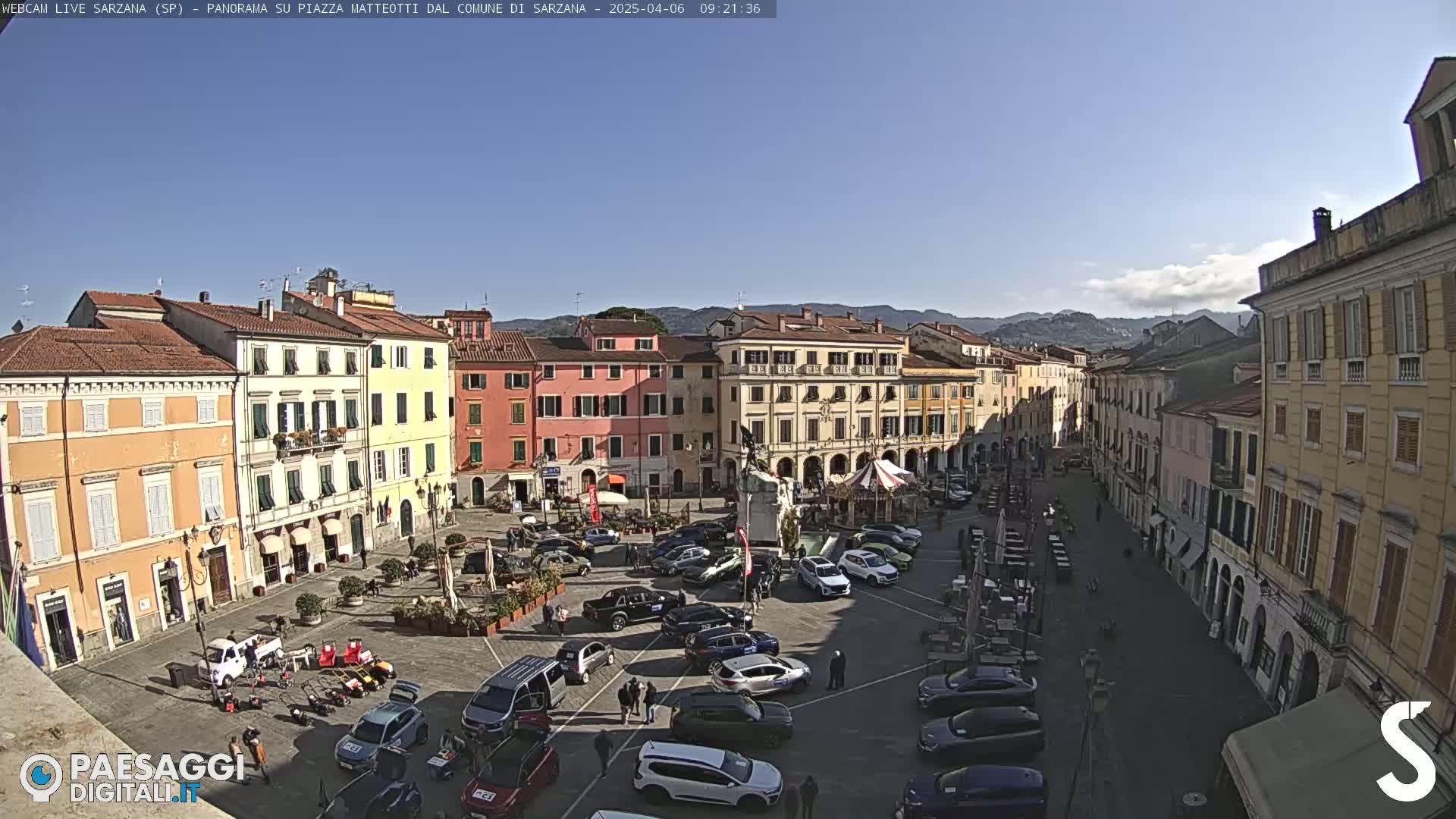 A sunny day in a town square with several parked cars and buildings surrounding the plaza.