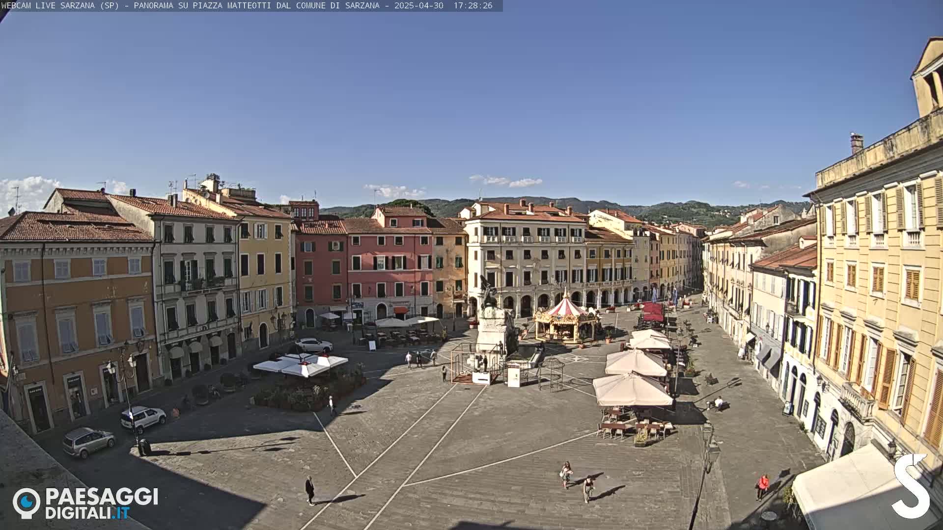 A mostly empty town square, under a clear blue sky, is surrounded by multi-story buildings.