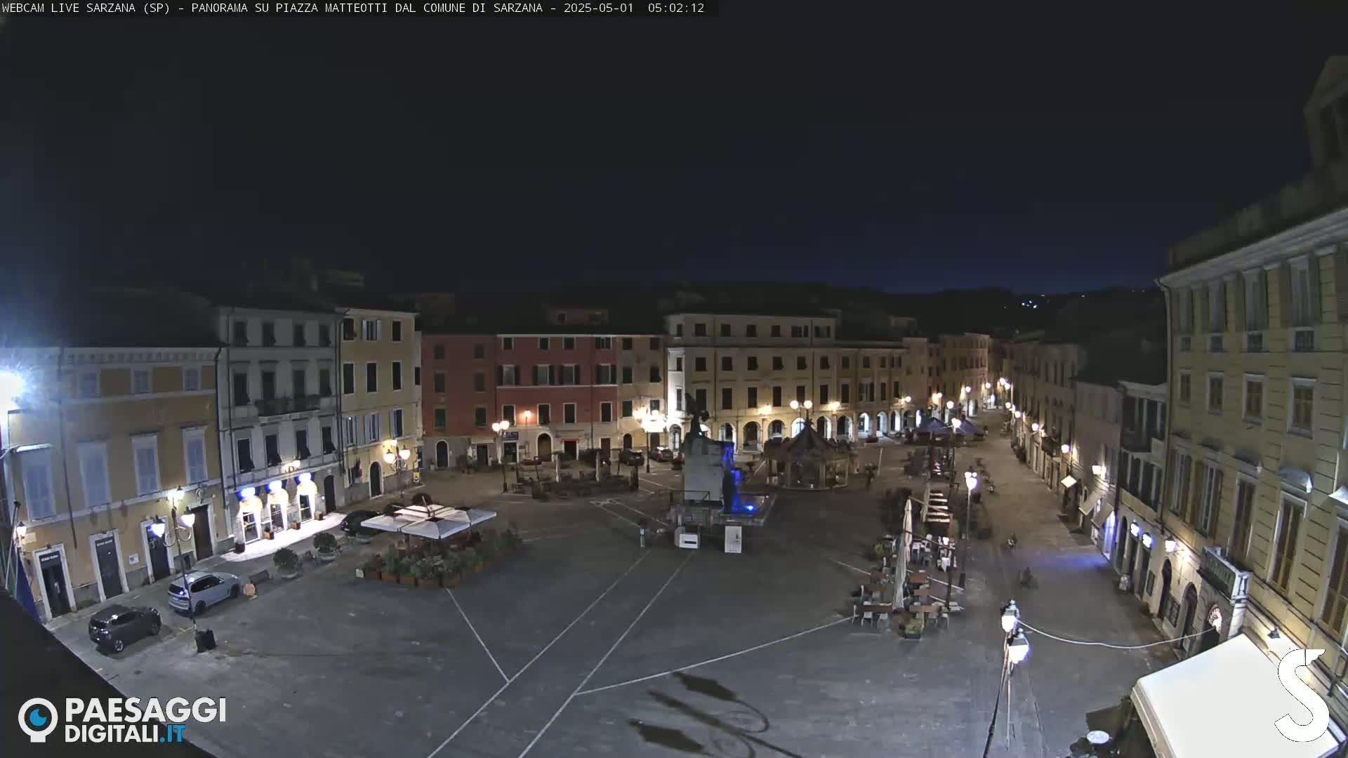 A nighttime, high-angle view of a town square with several buildings surrounding it, some with outdoor seating areas.