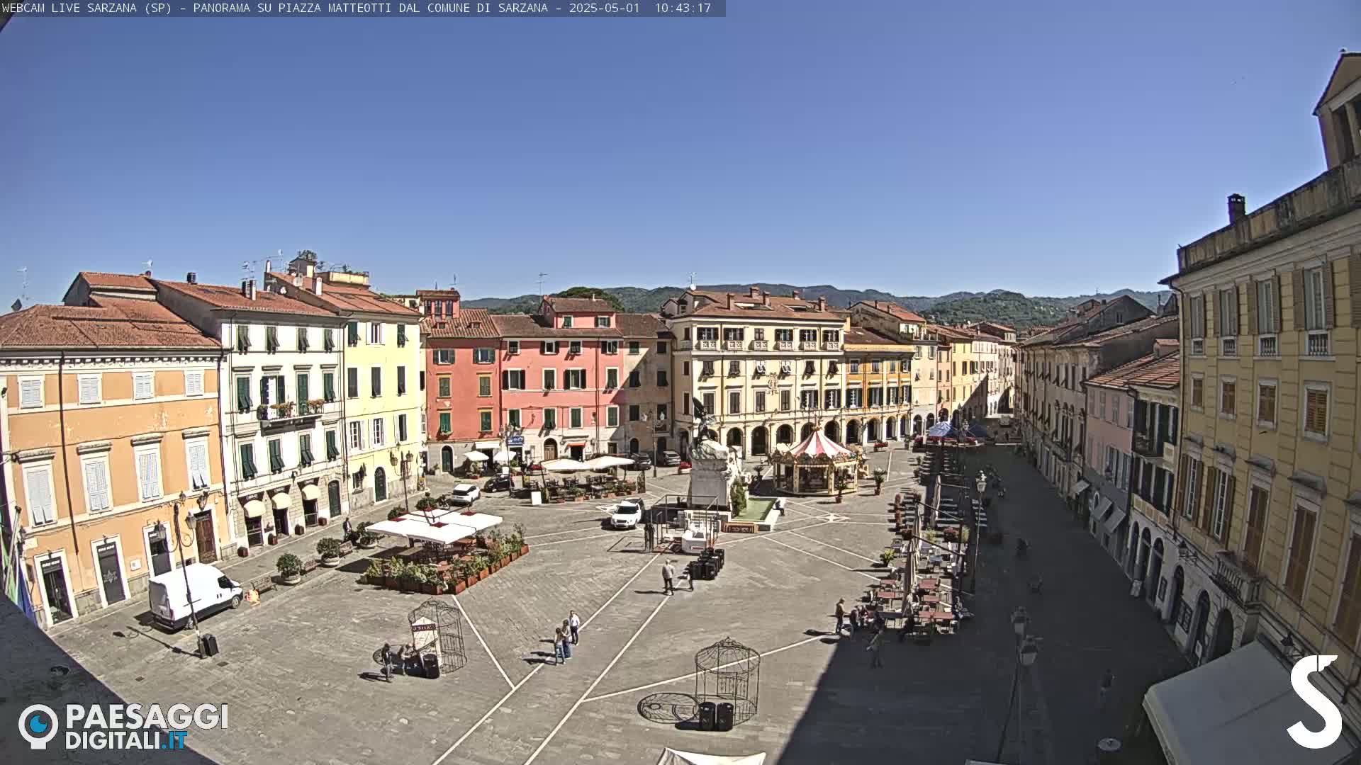 A large, sunny town square is surrounded by several multi-story buildings, with some outdoor seating areas and a few people walking around.