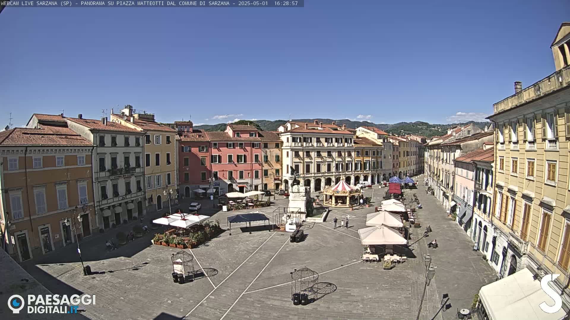 A sunny day shows a large town square surrounded by multi-story buildings, with a few people and outdoor seating areas visible.