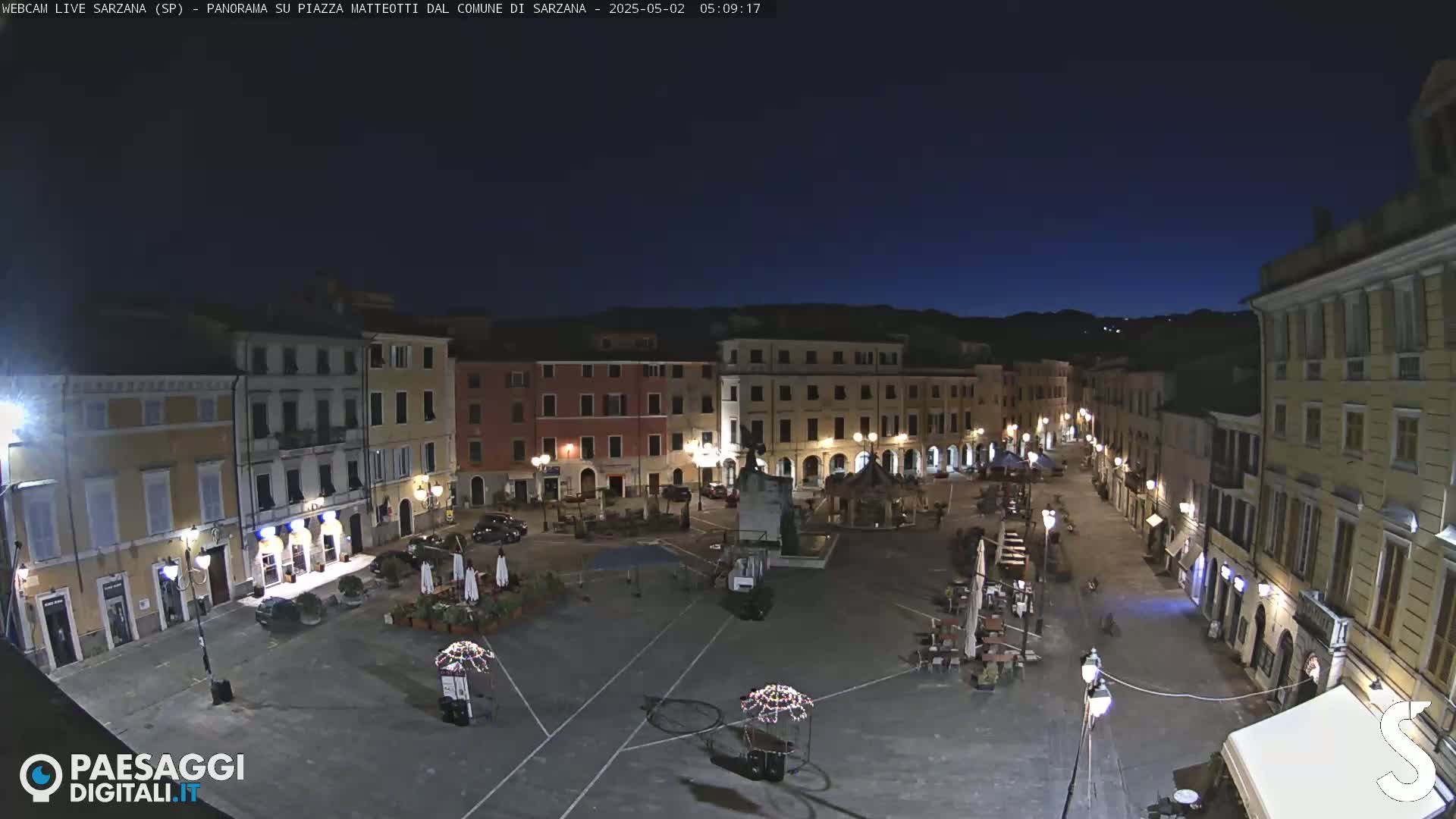 A nighttime view of a town square with several buildings surrounding it, under a dark blue, clear sky.