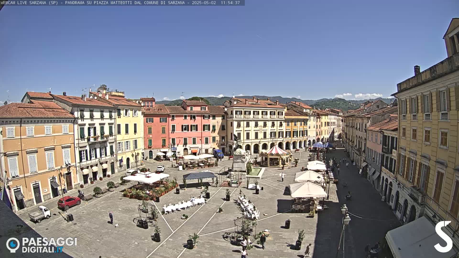 A sunny town square, surrounded by multi-story buildings, features outdoor seating and a carousel.