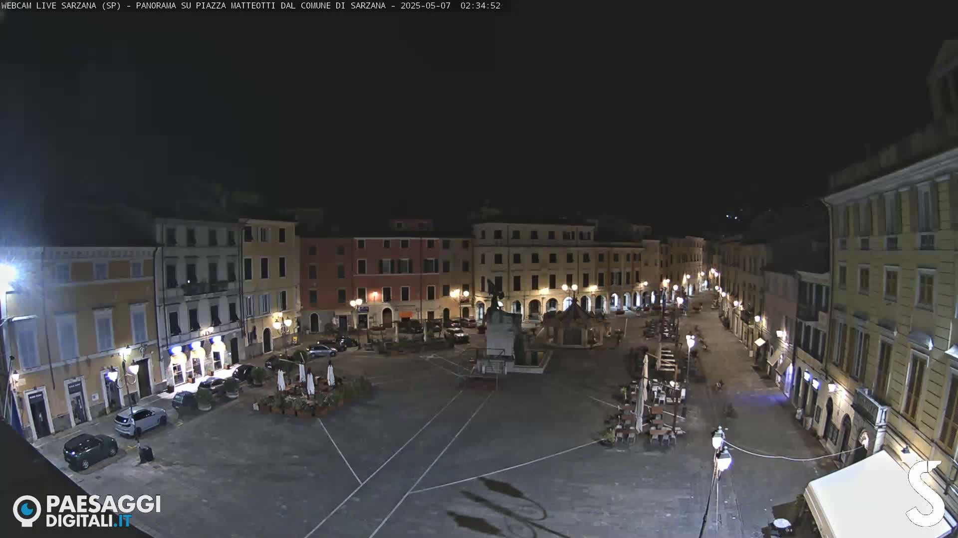 A nighttime, aerial view of a town square, surrounded by buildings with lights on, and a few cars parked in the square.