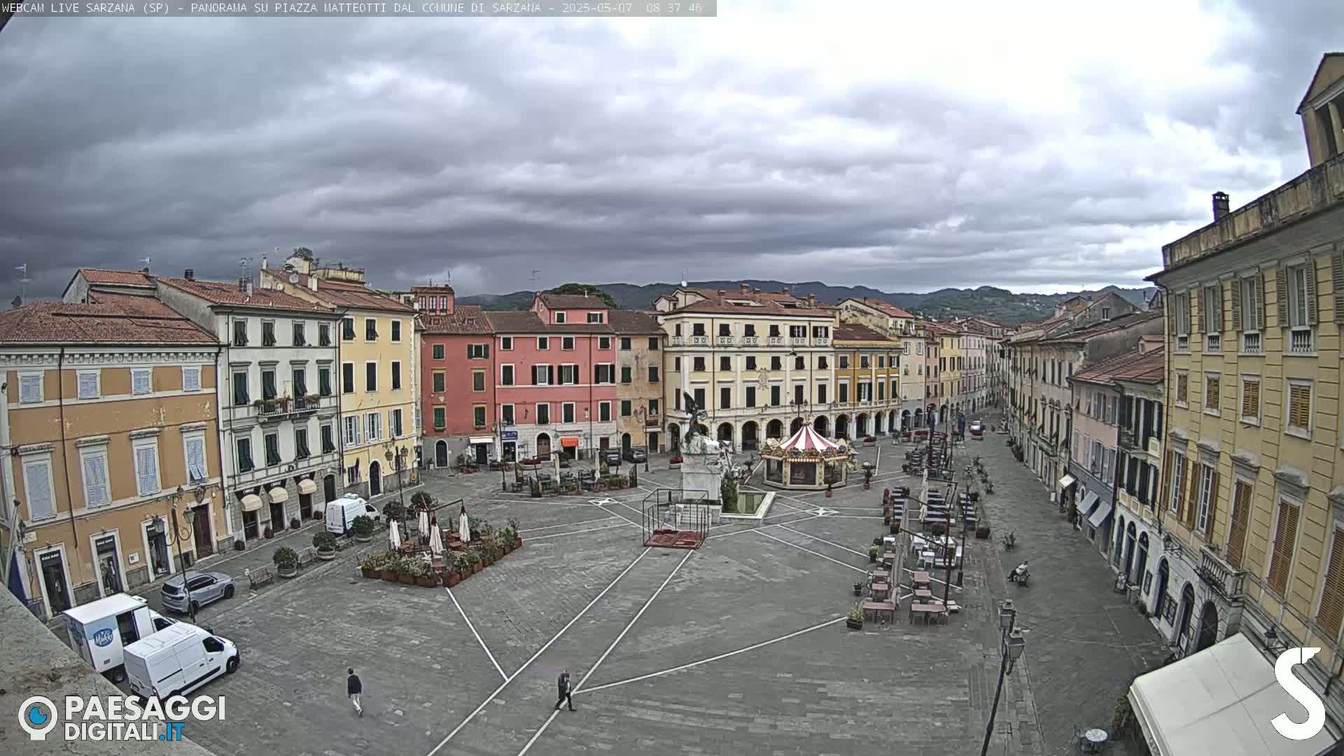 A mostly empty town square, surrounded by multi-story buildings, under a cloudy sky.