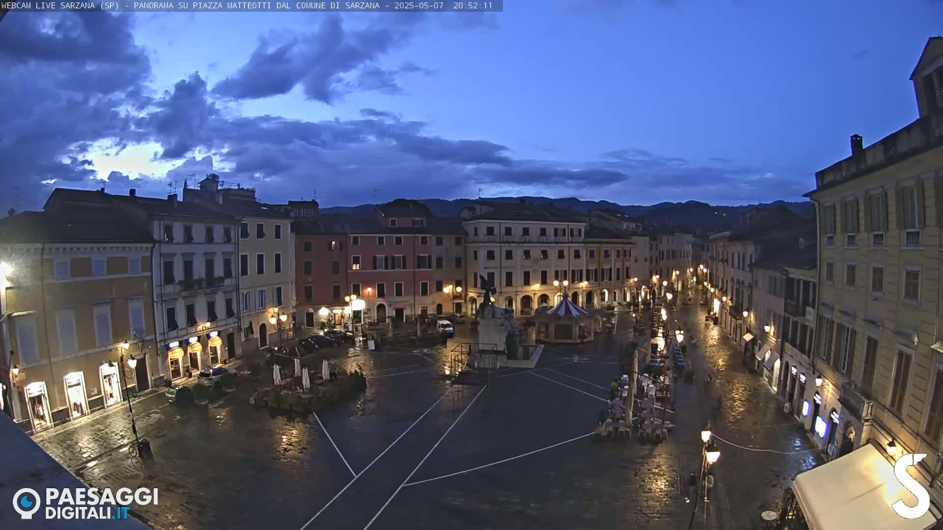 A wet, town square at twilight is surrounded by multi-story buildings with illuminated streetlights.