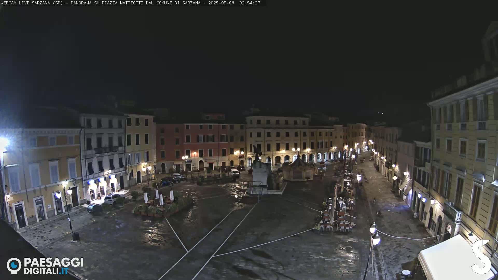 A wet, nighttime view of a town square with several buildings surrounding a central monument, sparsely populated with cars and outdoor seating.