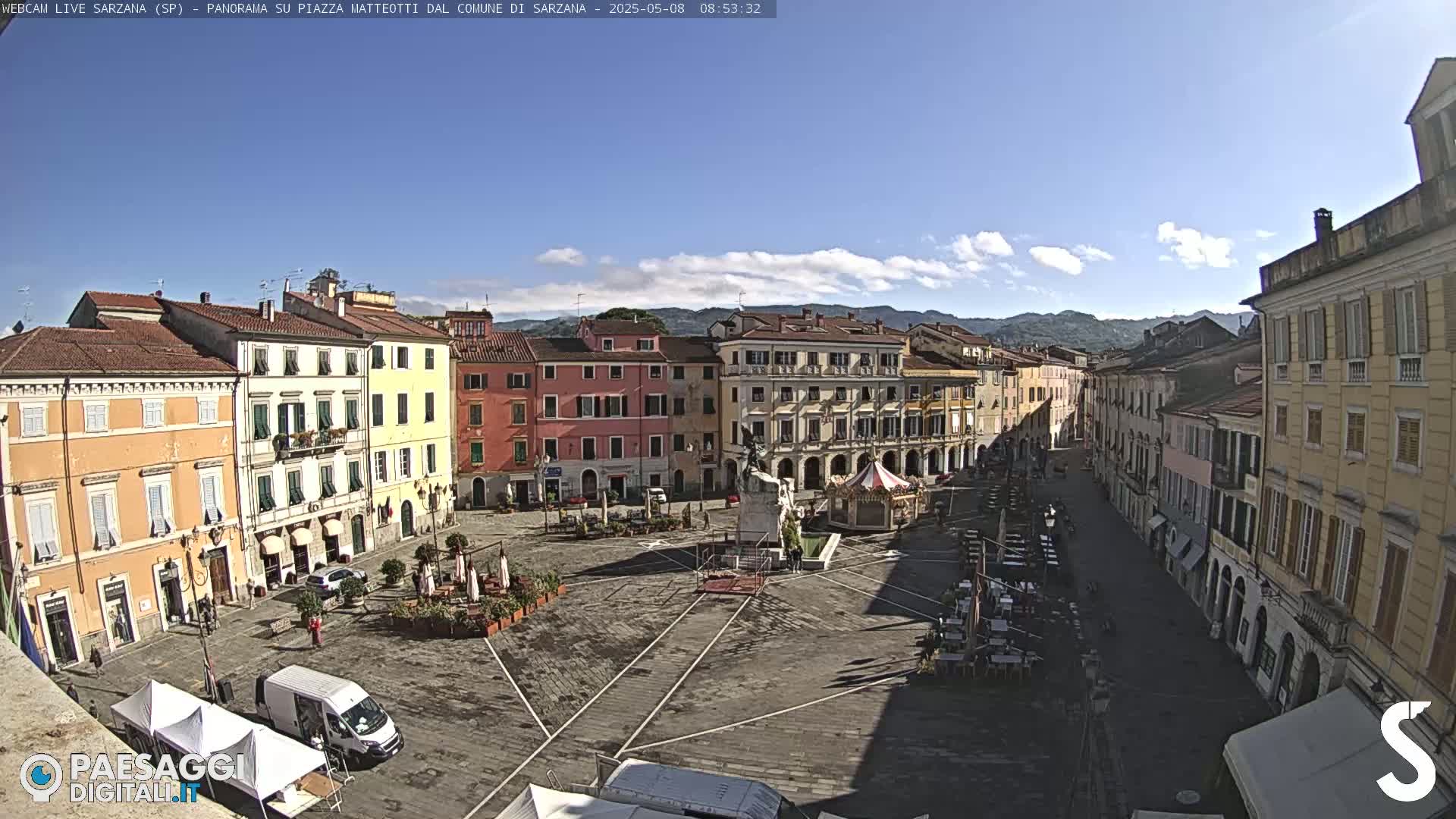 A mostly sunny day shows a large town square with several multi-story buildings surrounding it, a few outdoor cafes set up, and a monument in the center.