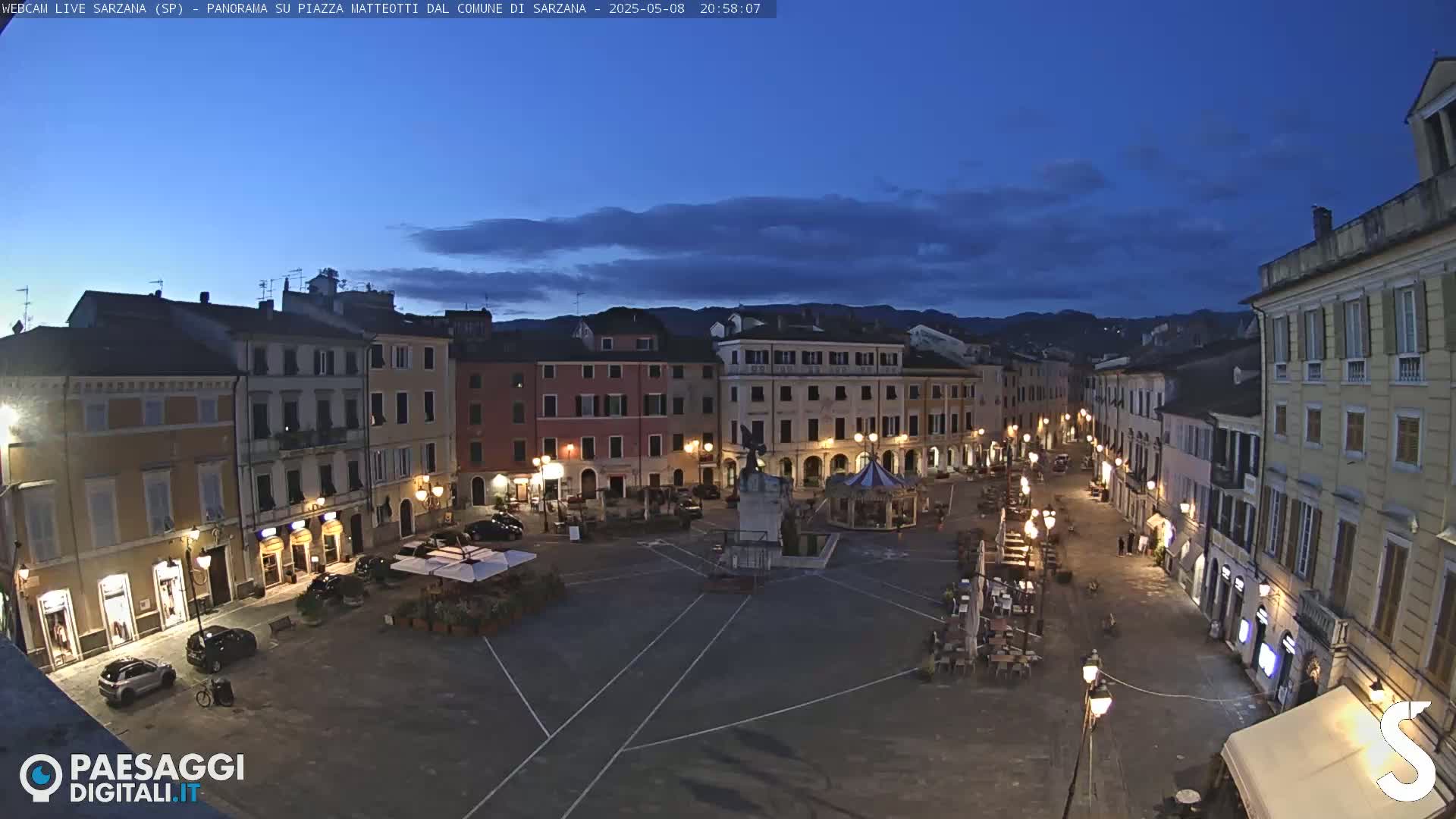 A town square at twilight, under a mostly clear, dark blue sky, is illuminated by streetlights, with cars parked along the edges and a few people in the distance.