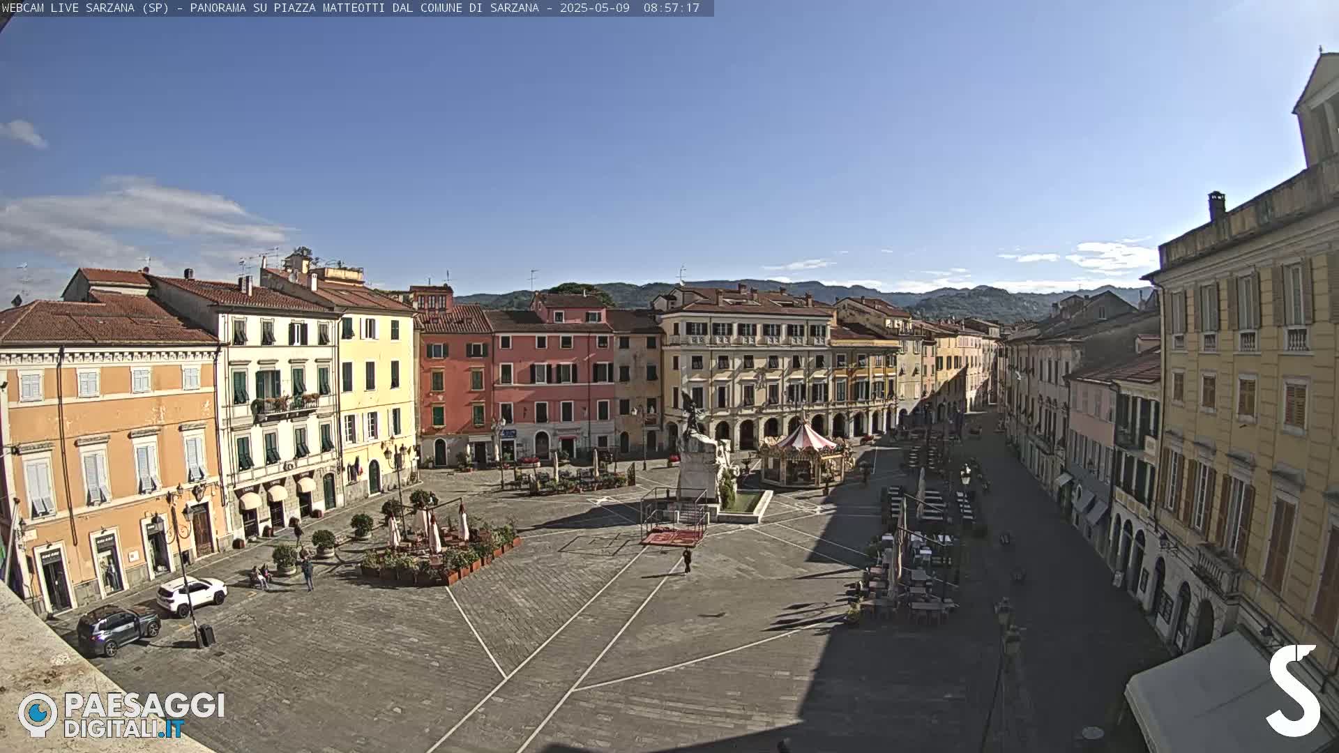 A sunny day reveals a large, paved town square surrounded by multi-story buildings, with a few people and some outdoor seating visible.
