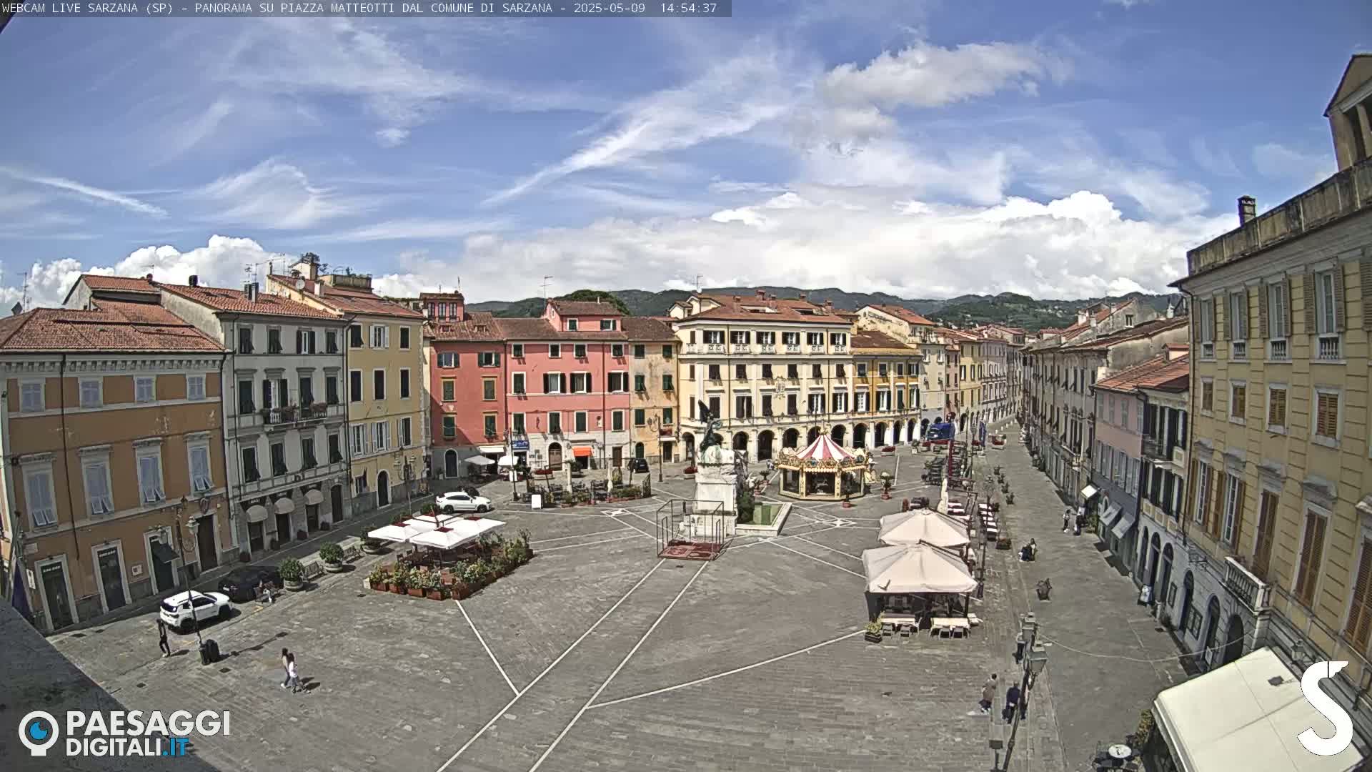 A mostly sunny town square with several multi-story buildings, a monument, a carousel, and outdoor seating areas.