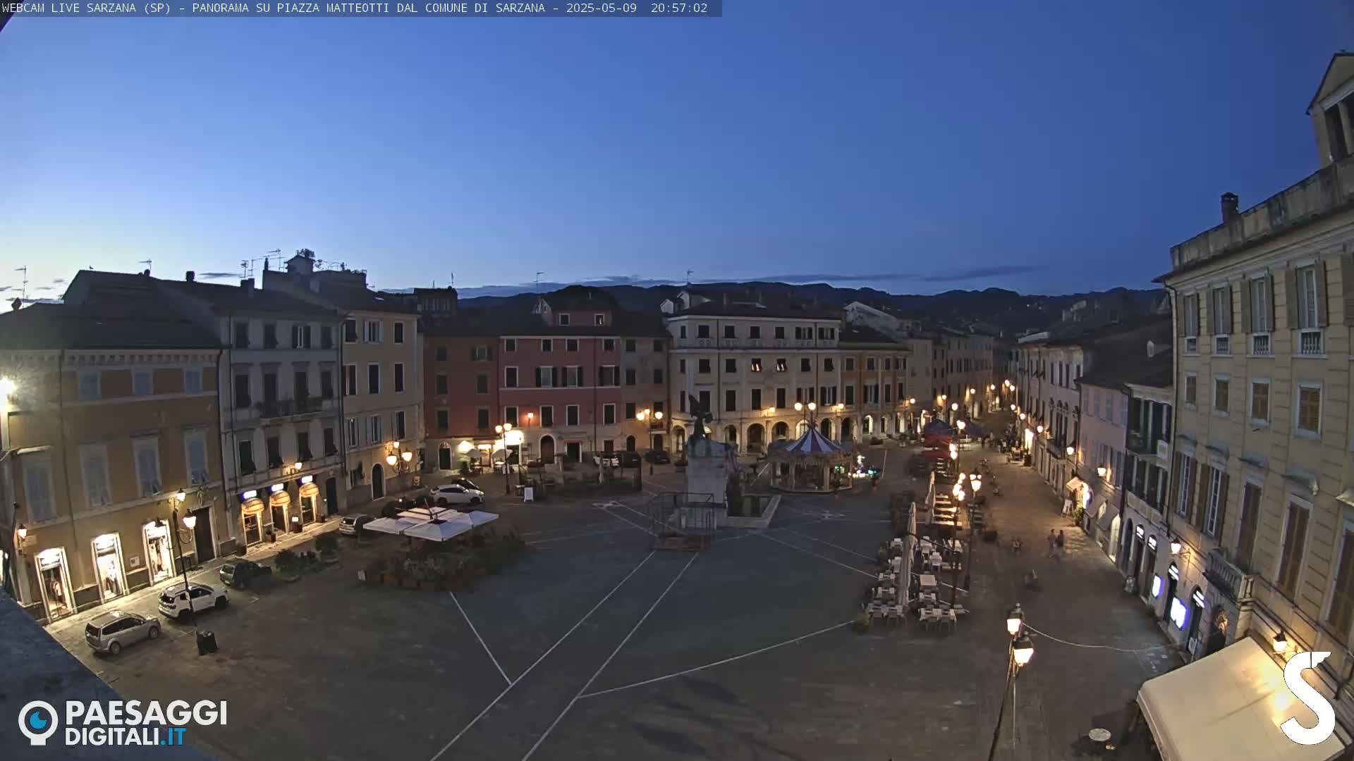 A twilight view of a town square, surrounded by multi-story buildings, with cars parked and outdoor seating areas in use.
