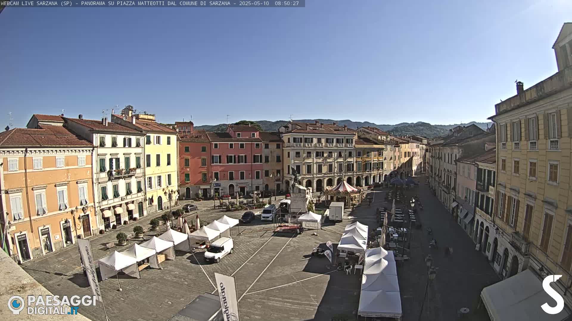 A sunny town square with several temporary market stalls set up, surrounded by multi-story buildings.