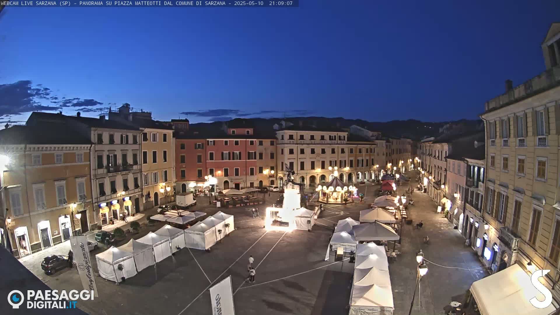 A town square at night, illuminated by streetlights, contains several white tents and a carousel, surrounded by multi-story buildings under a clear, dark blue sky.