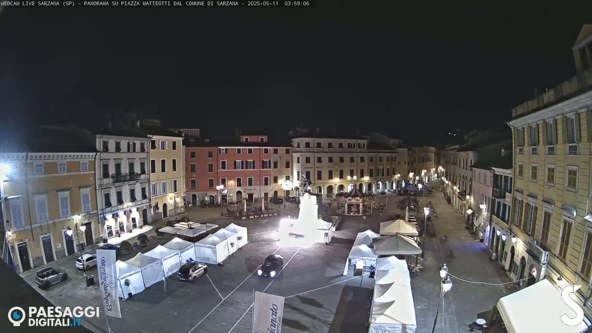 A nighttime view of a town square with several buildings surrounding it, illuminated by streetlights and containing numerous white tents, a monument, and a few cars.
