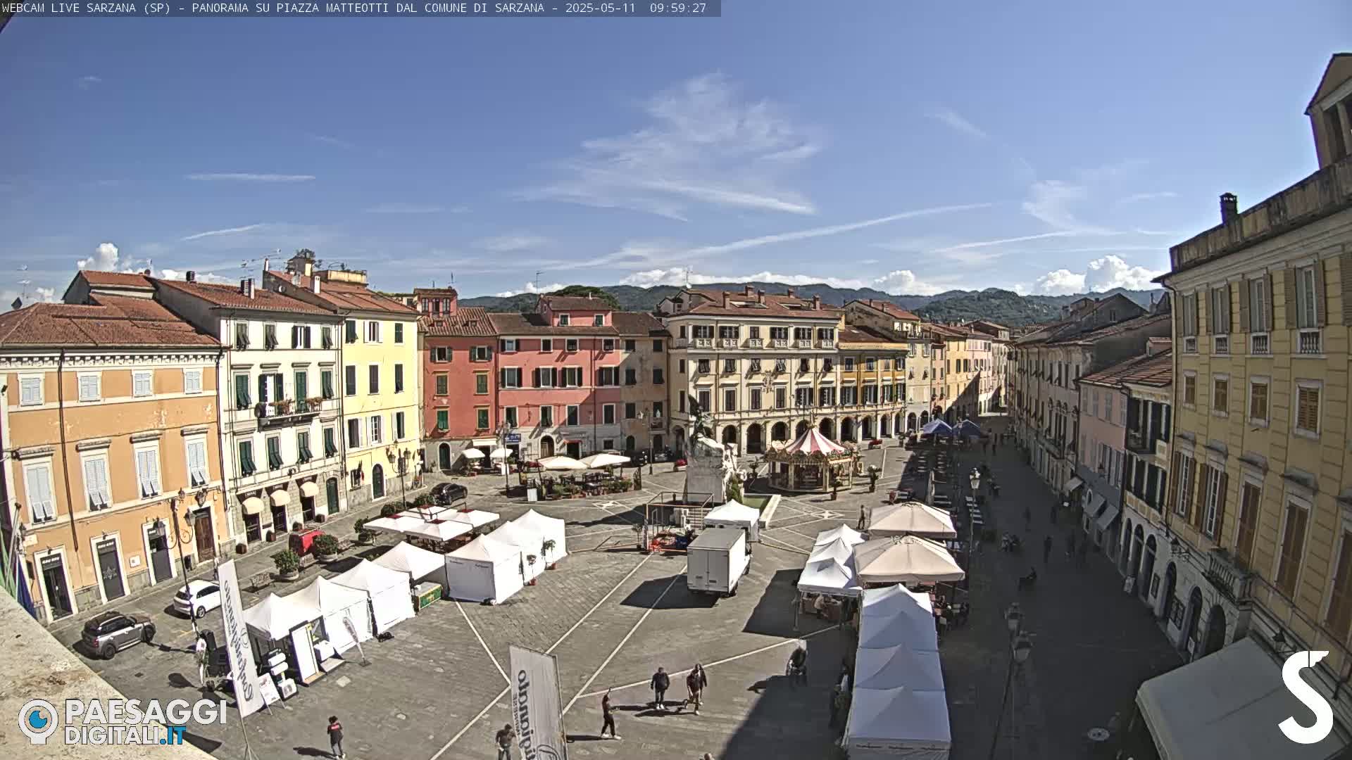 A sunny town square with several white tents, a carousel, and numerous buildings surrounds the square.