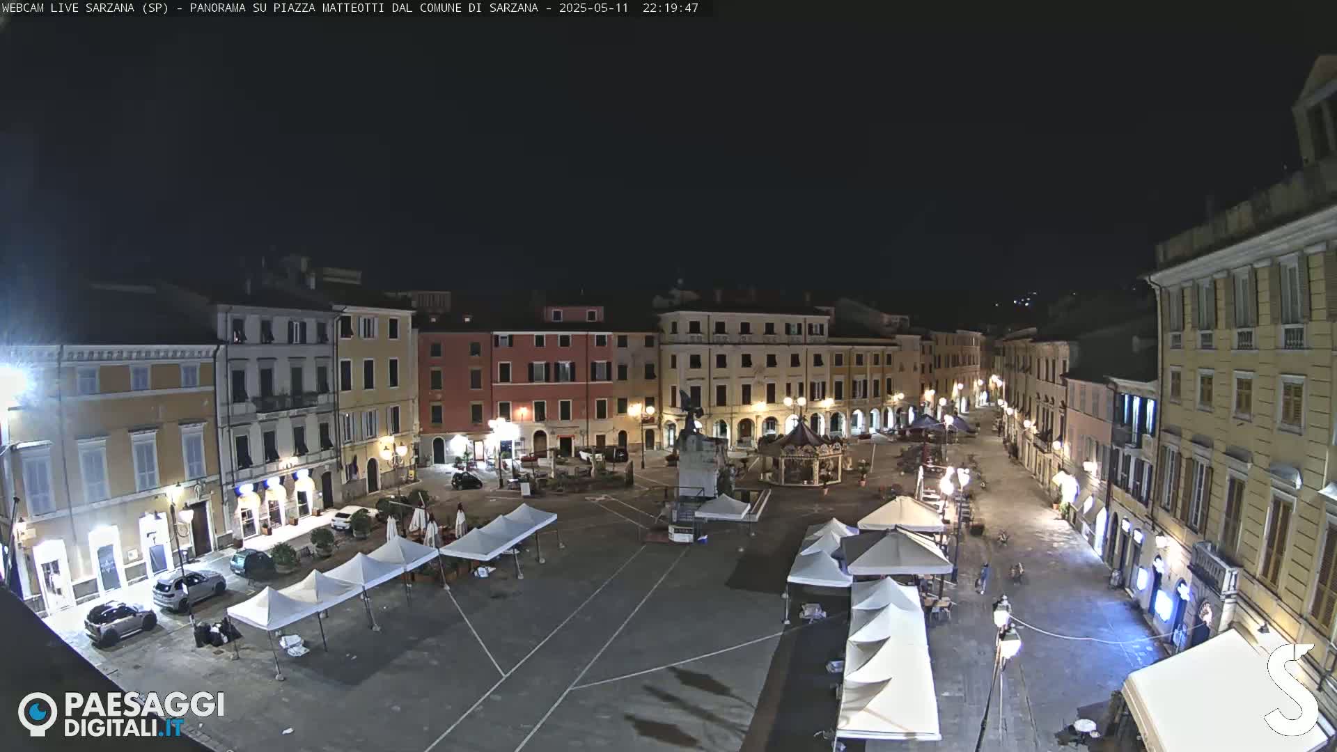 A nighttime view of a town square, with several buildings surrounding it, and numerous white canopies set up in the square.