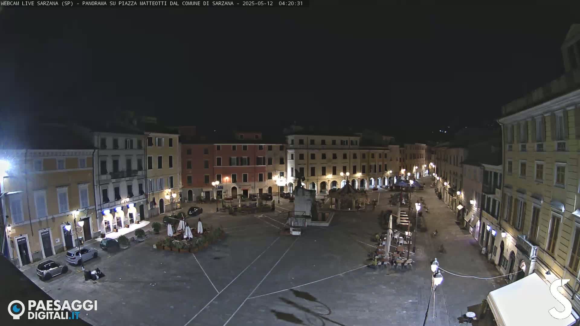 A nighttime view of a mostly empty town square surrounded by buildings, with a few cars and outdoor seating areas visible.