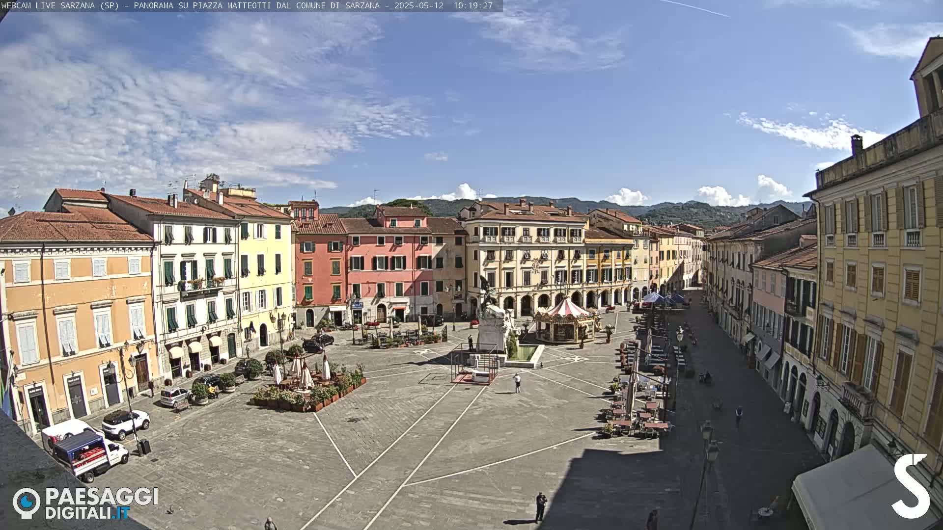 A sunny day reveals a large town square surrounded by multi-story buildings, a monument, and a carousel.