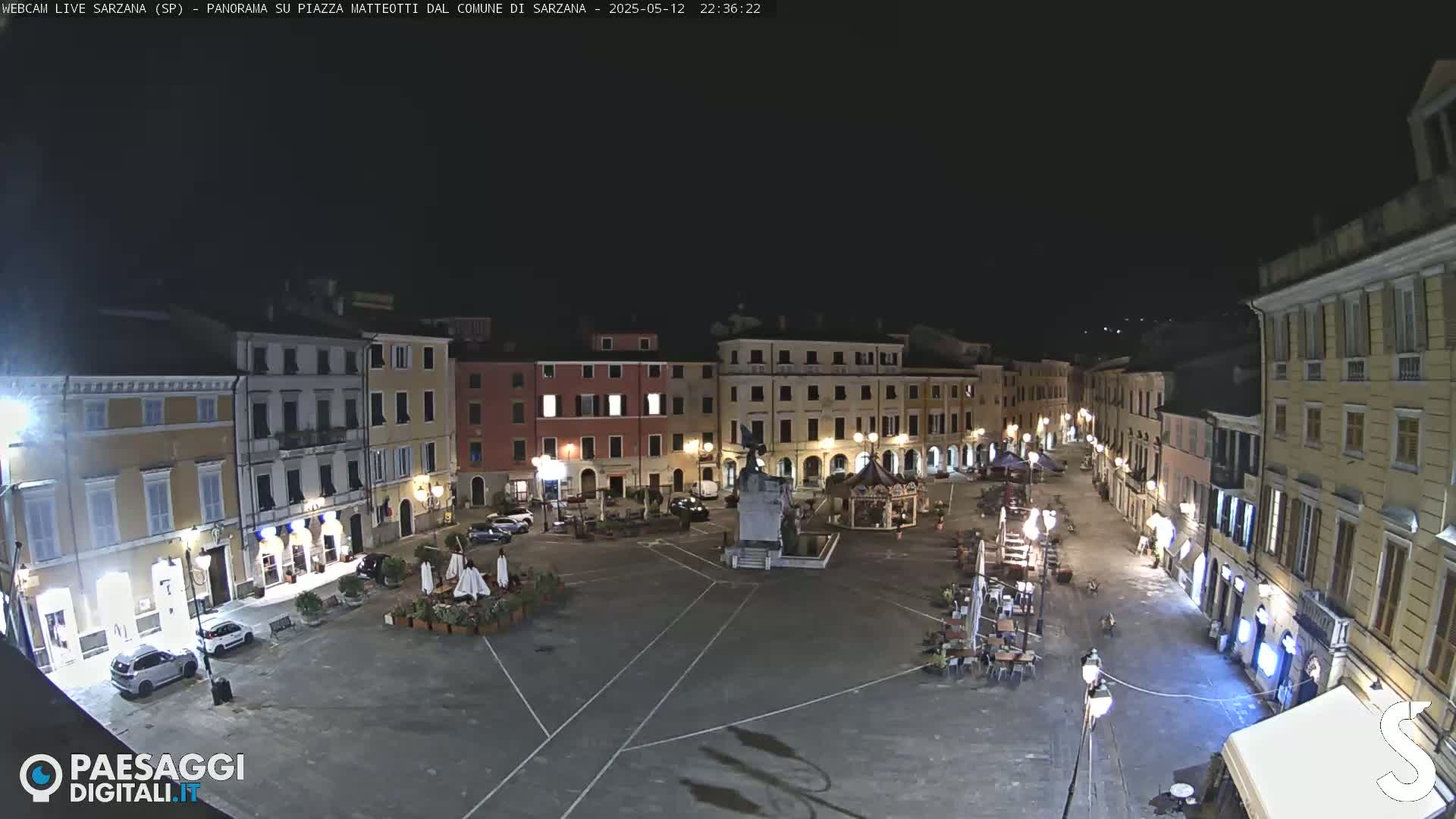 A nighttime, high-angle view of a town square with several buildings surrounding it, illuminated by streetlights, with a few cars parked and some outdoor seating visible.