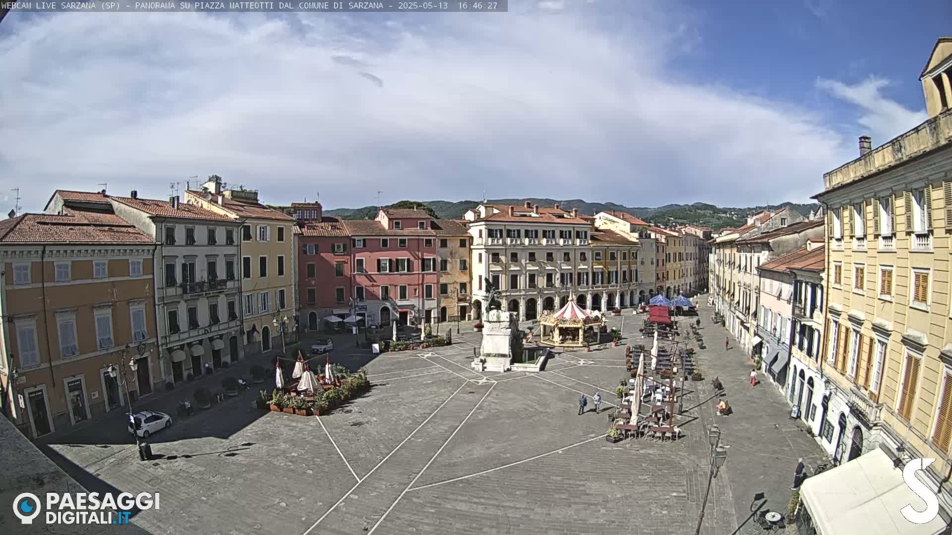 A mostly sunny day reveals a large, paved town square surrounded by multi-story buildings, with a few people and outdoor seating areas visible.