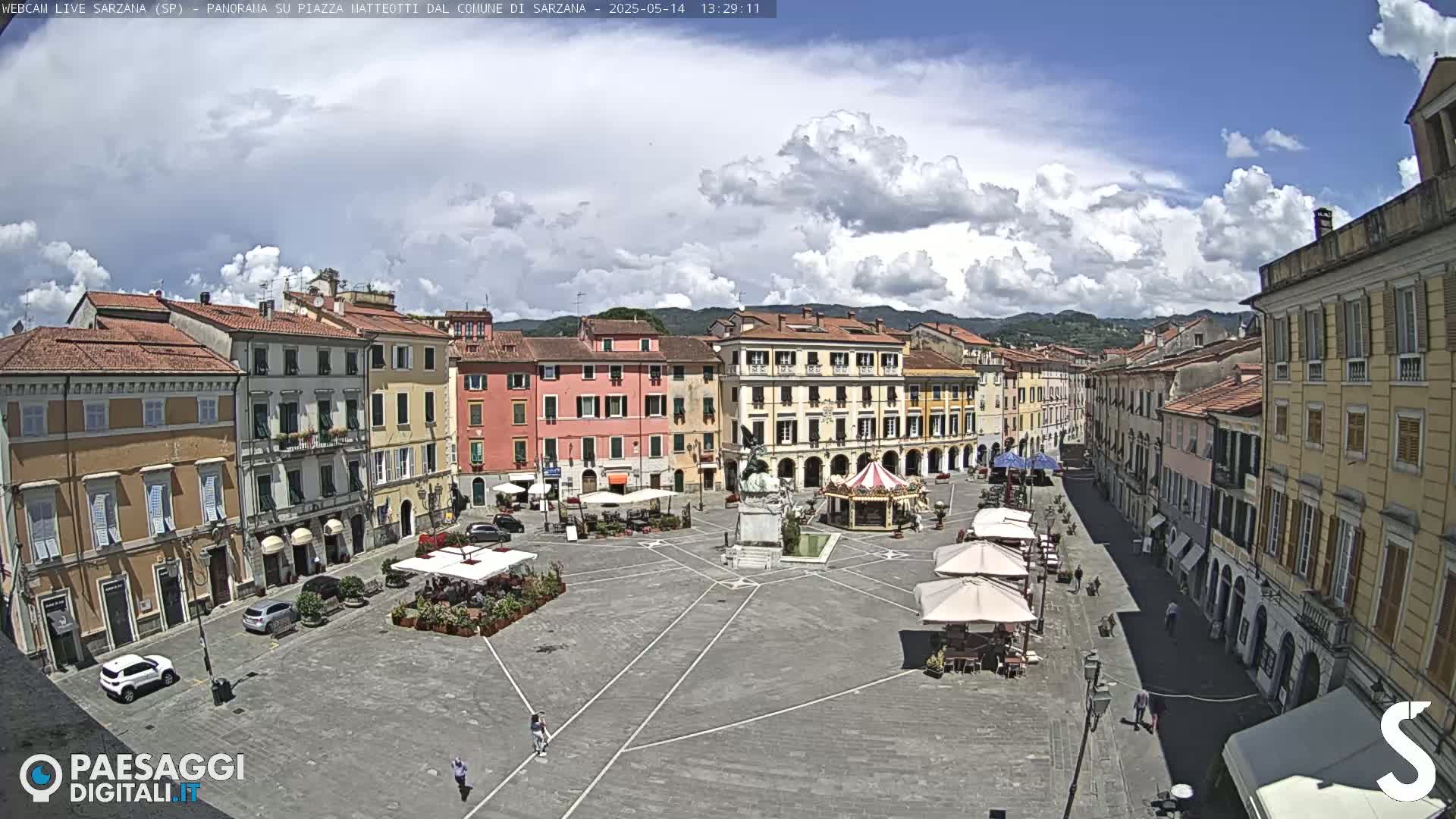 A partly cloudy day in a town square surrounded by multi-story buildings, with some people walking and several outdoor café umbrellas set up.