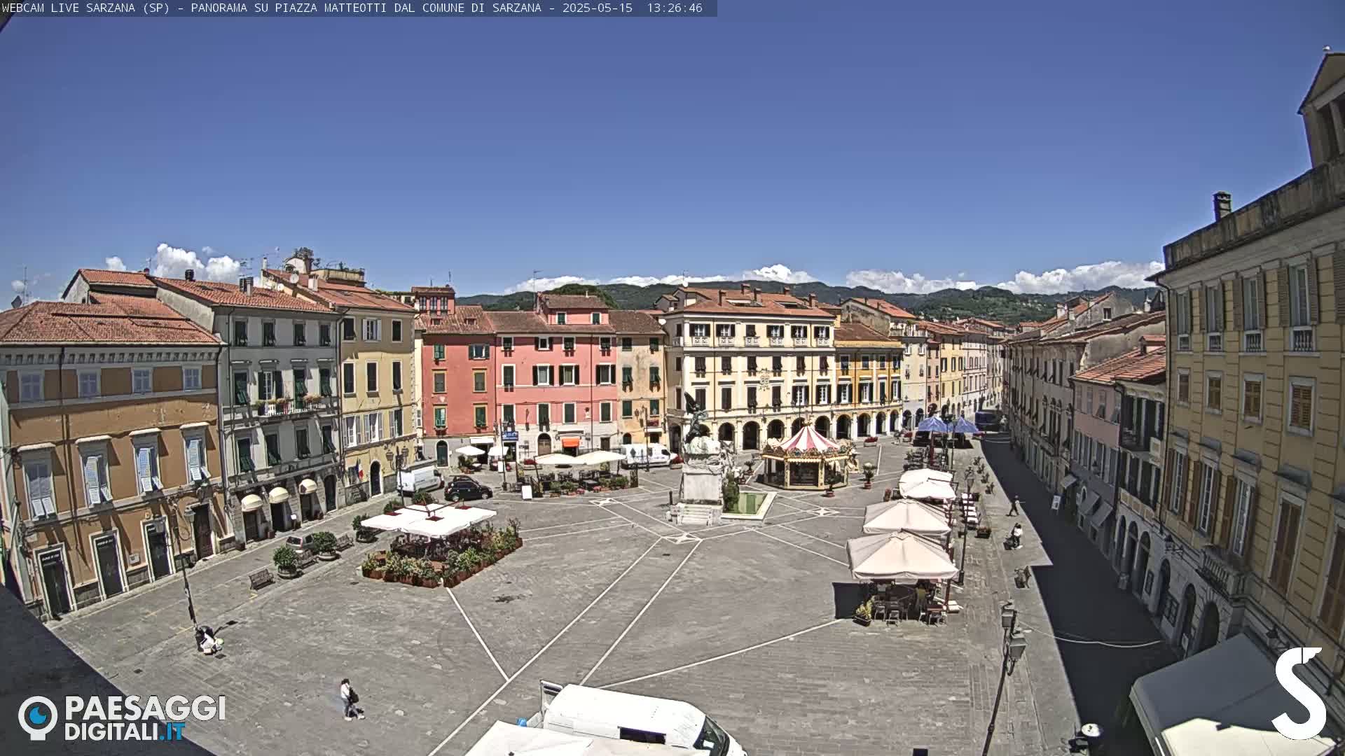 A mostly sunny, outdoor view of a large town square in Italy, surrounded by multi-story buildings, with several small outdoor cafes and a carousel.