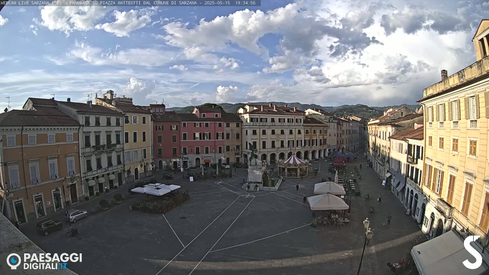 A mostly sunny town square with multi-story buildings surrounding it, a monument in the center, and a few people and outdoor seating areas.