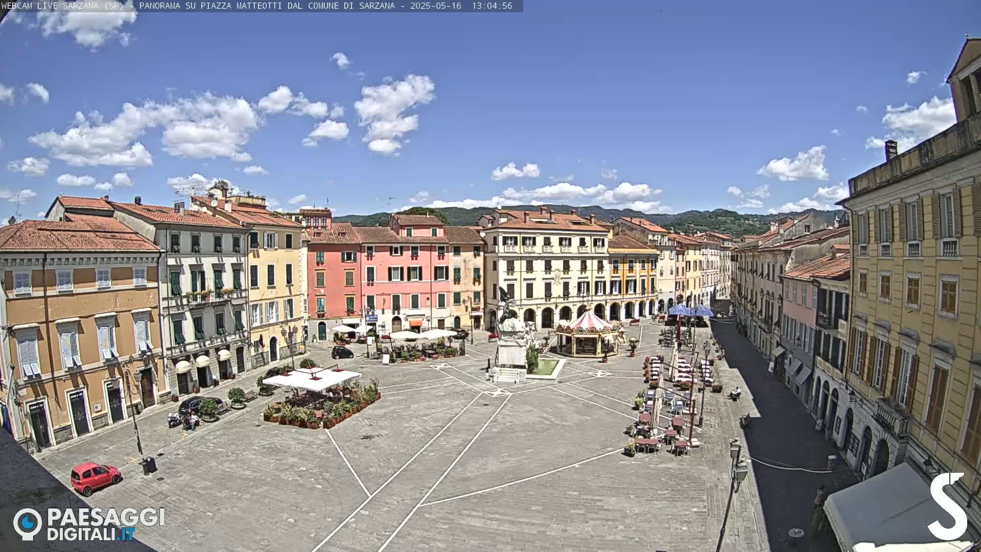 A mostly sunny day overlooks a large town square surrounded by buildings with a small carousel and outdoor seating.